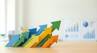 Colorful paper arrows pointing upward on wooden desk with blurred business charts and graphs in background representing growth, success and financial progress.