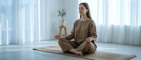 Young Caucasian woman in brown loungewear practicing meditation in lotus position on yoga mat in bright minimalist room with natural light.