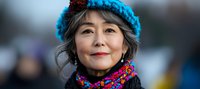Mature Asian woman wearing colorful winter knit hat and scarf smiling warmly at camera, expressing joy and contentment in cold weather outdoor setting.