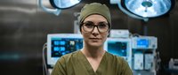 Female surgeon in green scrubs and cap standing confidently in modern operating room with surgical lights and medical equipment in background.