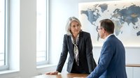 Professional blonde businesswoman in dark suit discussing strategy with male colleague in modern office with world map background for corporate presentations.