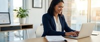 Professional Asian businesswoman working on laptop in modern office with natural sunlight streaming through windows creating productive workspace atmosphere.
