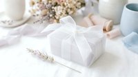 Elegant white gift box tied with sheer ribbon bow, lavender sprig beside it, soft floral decor in background, pastel wedding or bridal styling.
