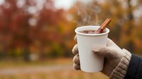 Gloved hand holding white paper cup of hot beverage with cinnamon stick, autumn foliage background. Seasonal drink concept for fall marketing and holiday promotions.