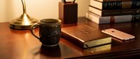 Cozy wooden desk with ceramic coffee mug, leather journal, smartphone, and stacked books in warm ambient light, perfect for productivity concepts.