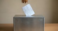 Hand placing ballot paper into transparent voting box during democratic election process on wooden table against neutral background.