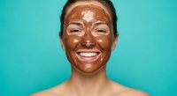 Happy young woman with chocolate face mask smiling against turquoise background. Skincare beauty treatment concept for spa and wellness routine.