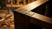 Traditional wooden joinery detail showing mortise and tenon connection in craftsman workshop with hand tools and lumber storage in background.