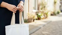 Person in dark clothing holding white canvas tote bag outdoors. Unrecognizable female model demonstrating reusable shopping bag for retail, eco-friendly lifestyle, and sustainable consumer products.