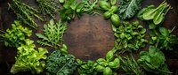 Fresh green herbs and leafy vegetables arranged in circular frame on rustic wooden background for healthy cooking and culinary design projects.