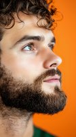 Profile portrait of young Caucasian male with curly hair and full beard looking up against vibrant orange background, showing contemplative expression.