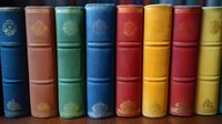 Row of vintage hardcover books with ornate gold embossed spines in various colors including blue, brown, green, red and yellow, arranged neatly on shelf showing classic binding.