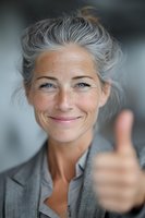 Confident mature woman with natural gray hair and freckles showing thumbs up gesture, expressing positivity and approval in professional setting.