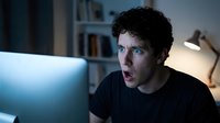 Young man with curly dark hair and light eyes, appearing shocked or surprised while working on laptop at night in home office setting with bookshelf and lamp.