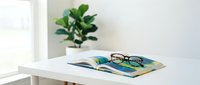 Eyeglasses and notebook on white desk with potted plant and window light, workspace organization and productivity concept.