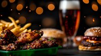 Juicy chicken wings, burgers and french fries on restaurant table with dark beer glass and festive bokeh lights in background. Appetizing pub food setup.