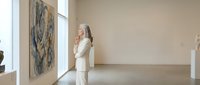 Elegant senior woman in white suit contemplating abstract artwork in modern minimalist gallery space with natural lighting and clean architectural lines.