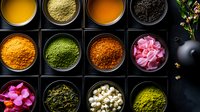 Colorful spices and tea ingredients in black square containers on dark background, including turmeric, matcha powder, rose petals, and jasmine pearls. Food photography.