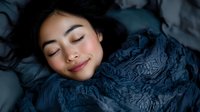 Young Asian woman sleeping peacefully with gentle smile on face, wrapped in dark blue textured blanket. Natural beauty and wellness concept for sleep articles.