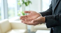 Professional businessman in dark suit with open cupped hands in protective gesture, symbolizing care, support and security in bright modern office environment.