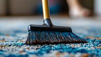 Close-up view of black broom sweeping blue carpet with scattered debris, showing detailed bristles and cleaning action in selective focus against blurred background.