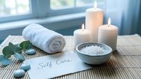 Spa self care setup with candles, sea salt, rolled towel, eucalyptus and stones on bamboo mat near window.