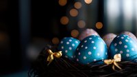 Blue polka dot Easter eggs decorated with golden bows nestled in dark nest against blurred bokeh lights background, creating festive spring holiday atmosphere.