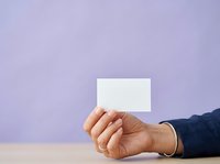 A person with medium skin tone holds a blank white business card or paper against a soft lavender background, ideal for corporate branding, marketing materials, and business presentations.