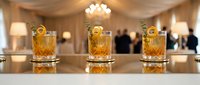 Three crystal whiskey glasses filled with amber liquor on elegant bar counter in upscale restaurant or hotel lounge with warm lighting and blurred guests.