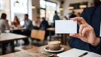 Professional holding blank white business card in modern office cafe setting with colleagues meeting in background, networking and corporate communication concept.