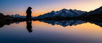 Silhouette of hiker enjoying peaceful mountain lake reflection at golden hour sunrise with snow-capped peaks creating perfect mirror image in calm water.