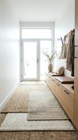 Bright, minimalist entryway with white walls, natural light from glass door, wooden storage bench, woven rugs, and hanging garments. Scandinavian interior design for home decor and lifestyle.