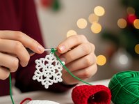 Woman hands holding white crocheted snowflake ornament with green yarn against festive Christmas lights bokeh background for holiday crafting projects.