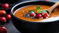 Creamy pumpkin soup garnished with fresh cranberries, basil leaves and black sesame seeds in black bowl on dark background, autumn comfort food.