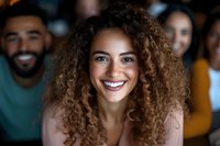 Young woman with curly brown hair and bright smile looking at camera, radiating joy and confidence in casual indoor setting. Natural lighting highlights genuine expression.