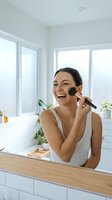 Happy young woman applying makeup with brush in bright modern bathroom mirror laughing joyfully during morning beauty routine skincare cosmetics.