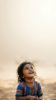 Young girl with dark curly hair and medium brown skin tone, wearing striped blue and red shirt, smiling upward outdoors at sunset or sunrise with soft blurred background.