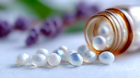 Small white homeopathic pills spilling from amber glass bottle on blurred purple floral background, alternative medicine and natural healing concept.