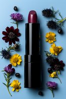 Burgundy lipstick surrounded by yellow daisies, purple chrysanthemums, dark blue thistles and black berries on light blue background. Flat lay cosmetic composition.