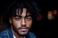 Young African American man with dreadlocks and facial hair wearing denim jacket against dark background with soft bokeh lights, expressing confidence and style.