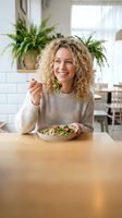 Happy young Caucasian woman with curly blonde hair eating healthy salad in modern kitchen with plants, smiling while enjoying nutritious meal at home.