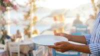Waiter in striped shirt holding empty white plate at outdoor rooftop venue with blurred city skyline and floral decorations in background.
