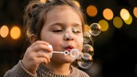 Young girl blowing soap bubbles with warm bokeh lights in background. Child enjoying playful moment with bubble wand creating magical atmosphere for childhood concepts.