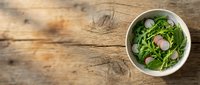 Fresh green salad with radish slices in white bowl on rustic wooden table, overhead view for food styling and culinary content.