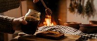 Cozy autumn evening with person pouring hot beverage into glass jar by fireplace, rustic wooden table with checkered cloth and dried herbs.