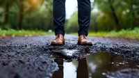 Man wearing brown leather oxford shoes and navy pants standing on wet asphalt road with puddle reflection after rain in autumn park setting.