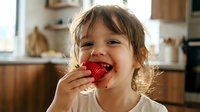 Happy toddler girl with light brown hair eating fresh red strawberry in bright kitchen, enjoying healthy fruit snack.