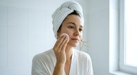 Young woman in white bathrobe and towel applying skincare cream with cotton pad in bright bathroom. Morning beauty routine and facial care concept.