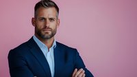 Confident businessman with beard wearing navy blue suit and light shirt standing with crossed arms against pink background, professional portrait.
