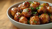 Glazed meatballs garnished with fresh herbs in white ceramic bowl on wooden table, appetizing close up view with selective focus and warm lighting.